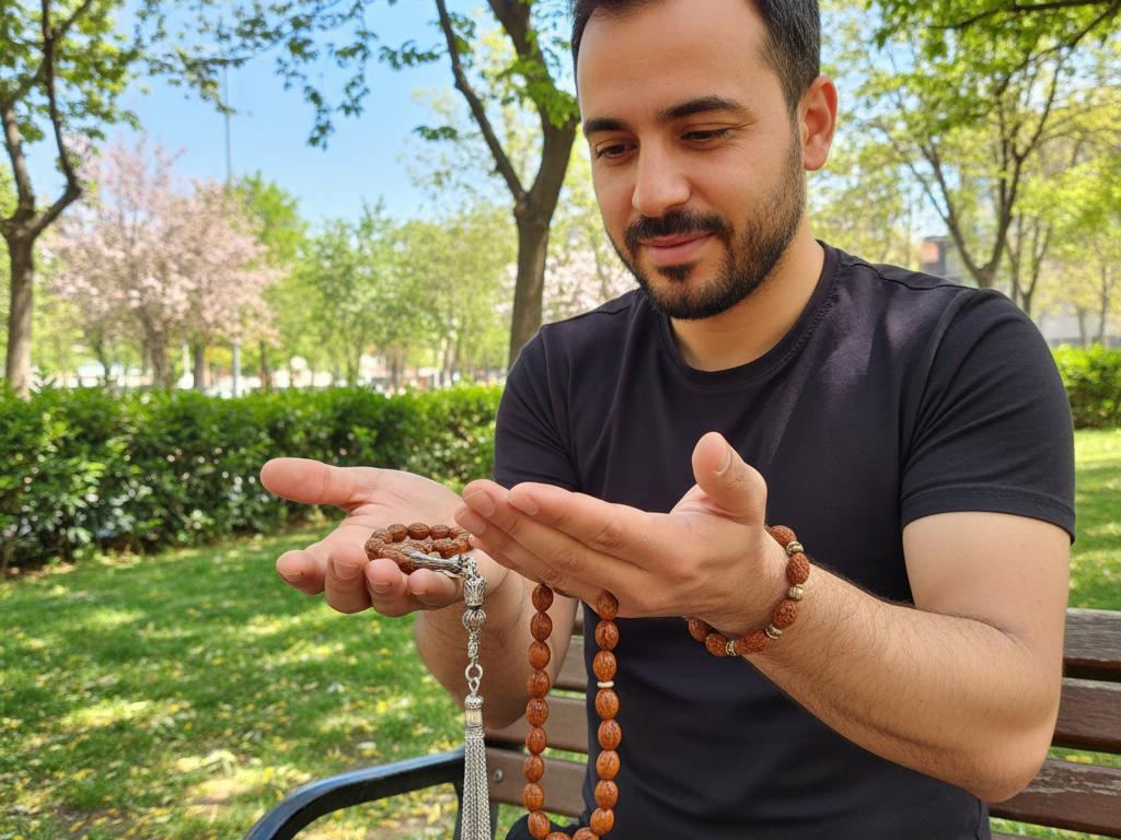 Photo of a middle-aged Turkish person outdoors in a sunny park holding and admiring a handmade tesbih and bracelet crafted from olive pits and İğde seeds, with green trees and blue sky softly blurred in the background.